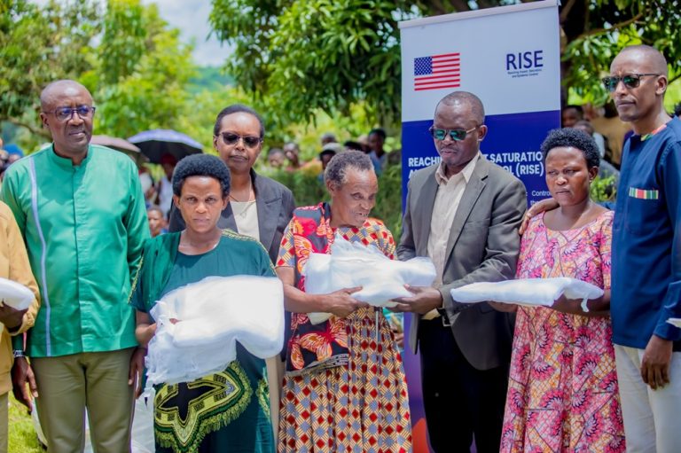 People outdoors at a blanket distribution event, receiving white bundles as a banner with the US flag and RISE logo looms in the background.