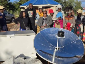 Group of people gathered outdoors around a large polished solar cooker dish and a chest freezer, watching or listening attentively.