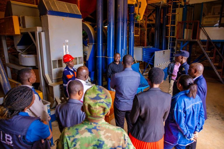 Group of workers and a presenter in an industrial facility listening to a briefing beside large blue machinery, with PPE like hard hats and vests visible regal in foreground.