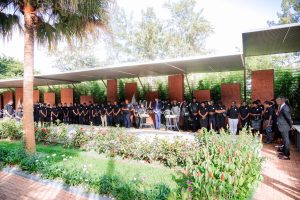 Outdoor memorial service with many attendees in black, standing under a covered stage as a speaker talks at the podium.