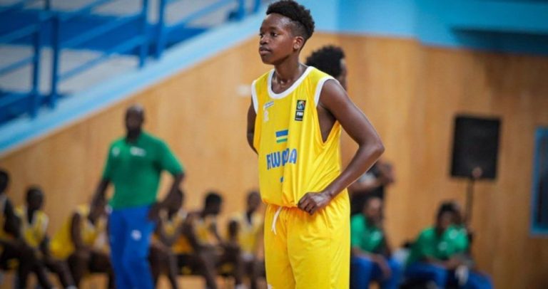 Young male basketball player in a yellow jersey stands with hands on hips on a gym court, teammates blurred in the background.