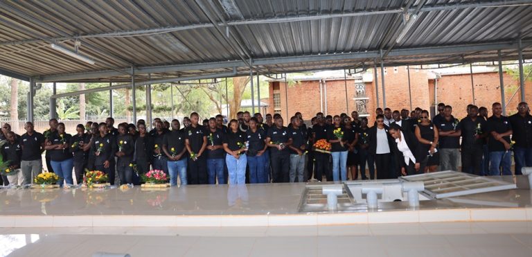 Large group of people in black stand under a metal-roofed shelter at a memorial service, holding flowers.