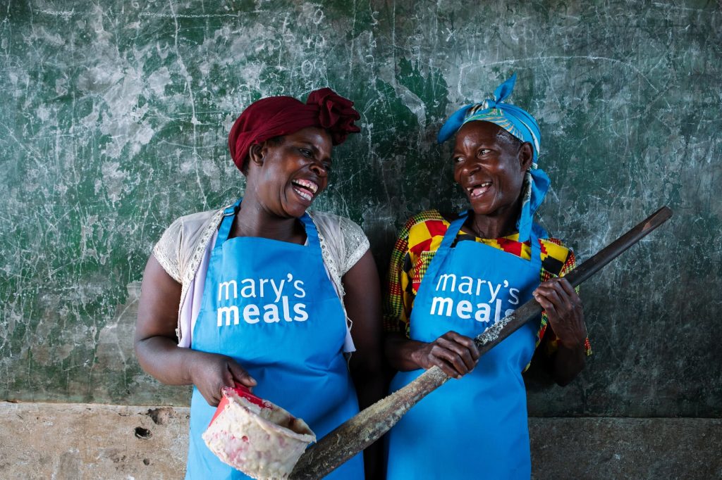 Two smiling women in blue aprons labeled 'Mary's Meals' stand together in a kitchen; one holds a meat cleaver with a piece of meat.