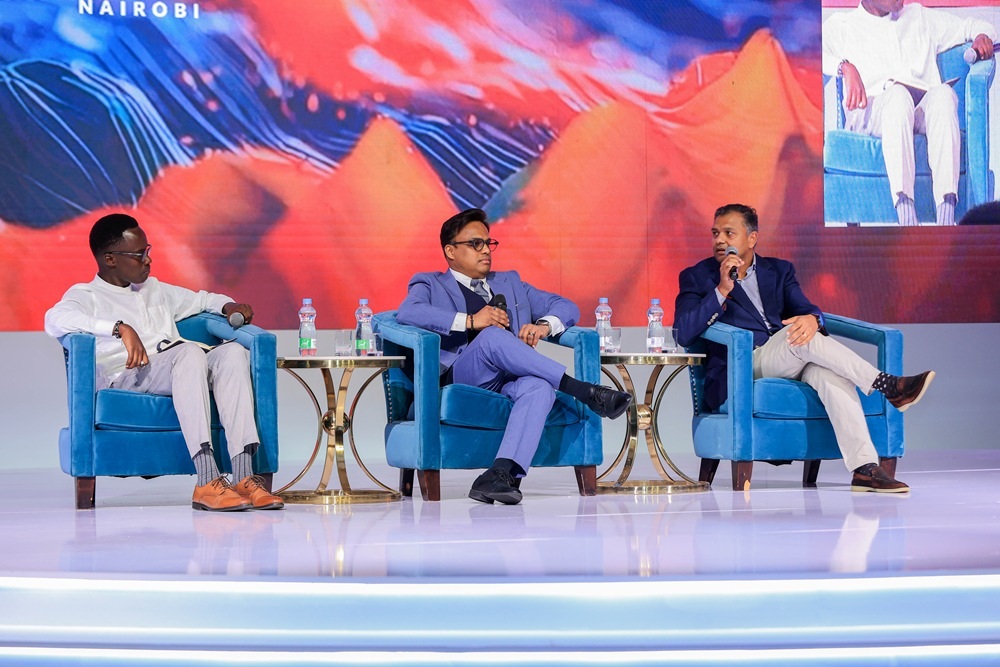 Three men seated in blue armchairs on a brightly lit stage, microphones in hand, speaking at a conference with a colorful backdrop.