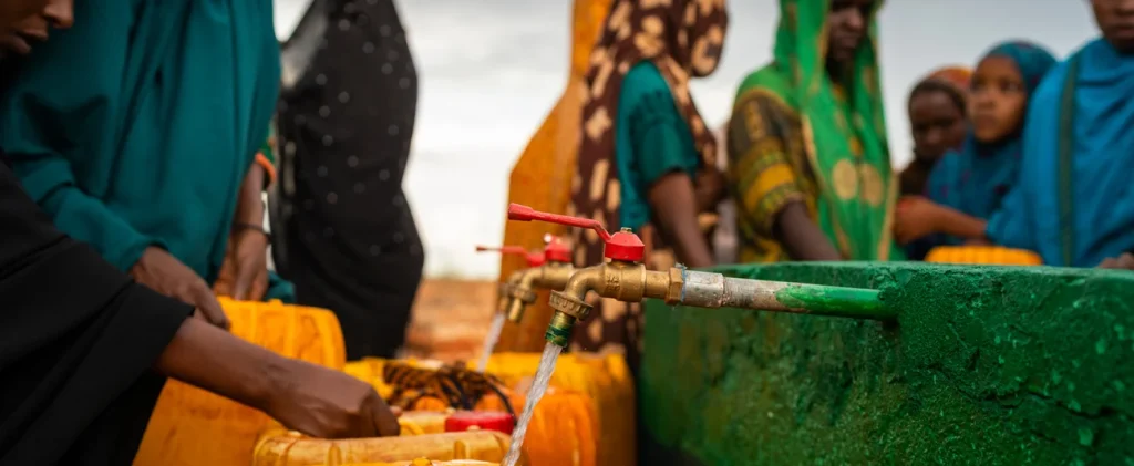 People in colorful traditional clothing fill yellow jerry cans with water from a communal tap outdoors.