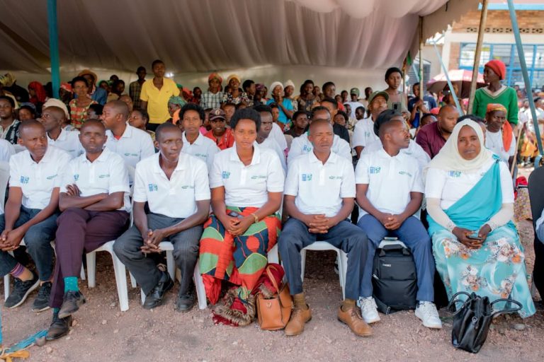 Group of people in white polo shirts seated in a row under a tent at an outdoor community event.