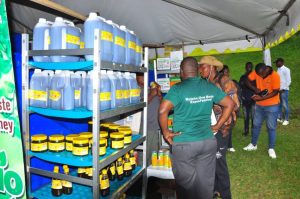 People gather around an outdoor expo booth under a tent, facing shelves stocked with blue jugs and yellow-labeled jars of product.