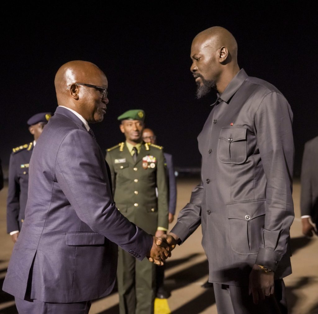 Two men in suits shake hands at night, with uniformed officers in the background.