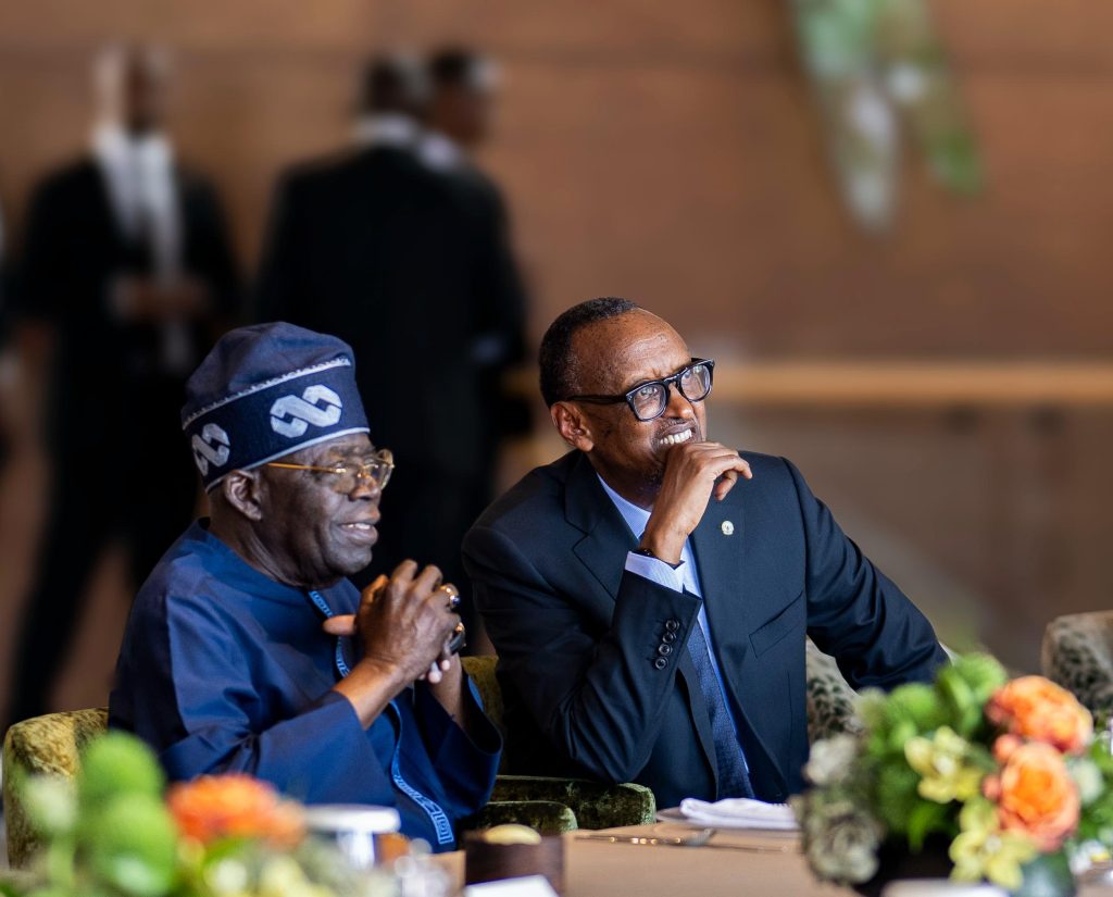 Two men in formal attire seated at a table during a ceremony; one in a suit with glasses, the other in blue traditional attire, flowers in front.