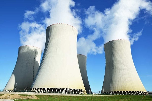 Three large concrete cooling towers emit steam against a bright blue sky at a power plant site with a green field in the foreground