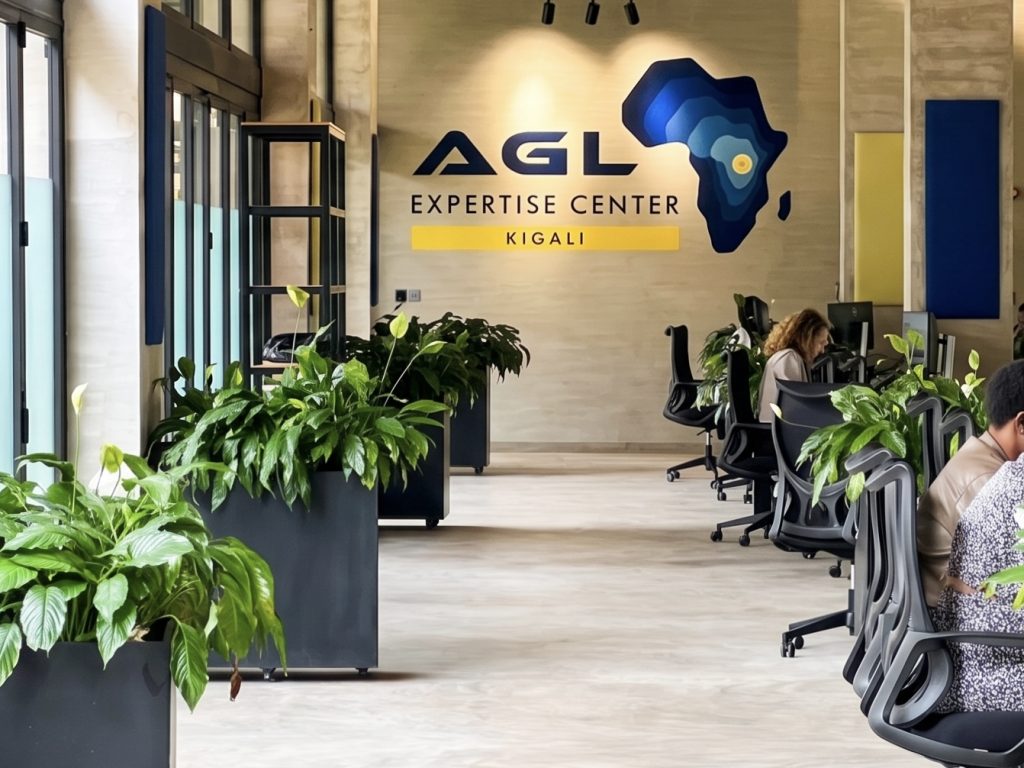 Office lobby with workstations, green leafy plants in tall planters, and a wall mural reading 'AGL Expertise Center Kigali' with a blue Africa-shaped graphic.—Informative image conveying location and setting.
