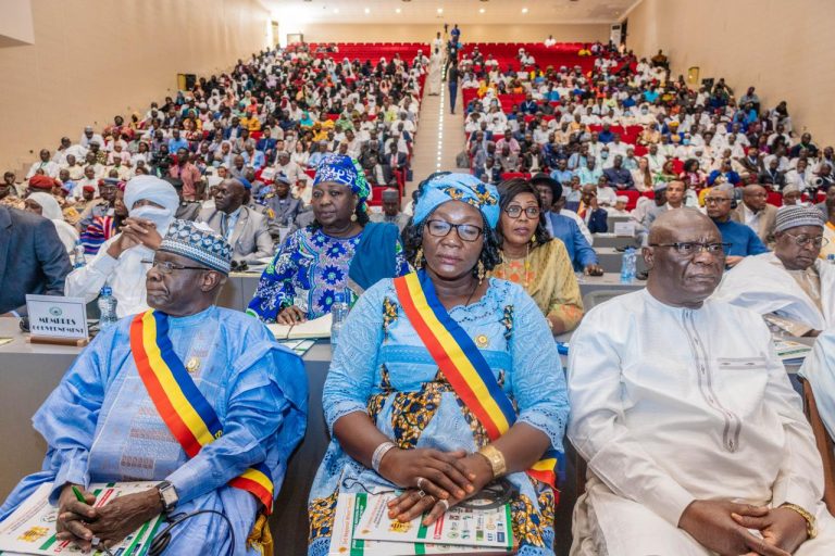 Front-row dignitaries in colorful traditional dress with tricolor sashes at a large conference; audience fills the auditorium behind them.