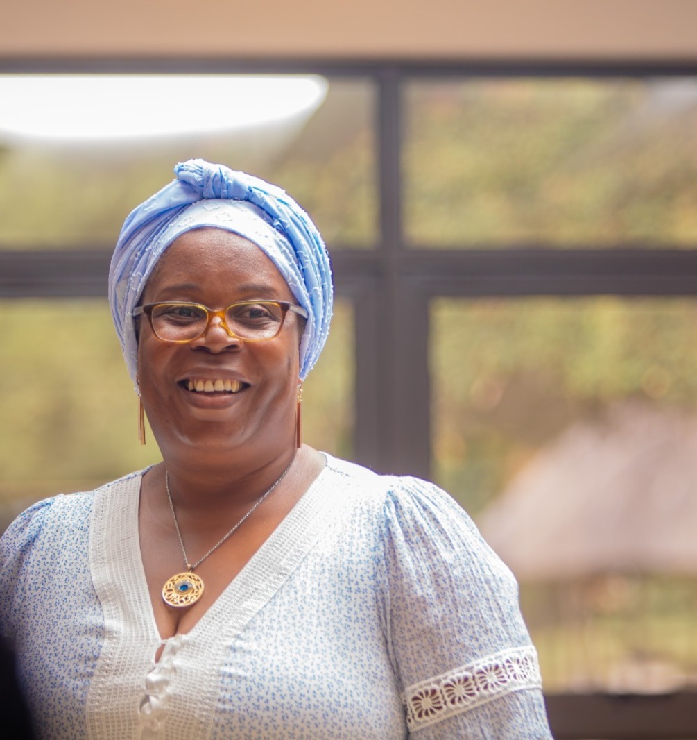 Smiling woman wearing a light blue head wrap and glasses, standing indoors with a soft backdrop behind her.