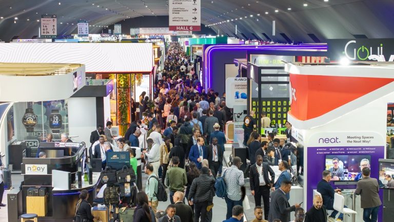 Busy trade show hall with colorful exhibition booths and a crowded central aisle filled with attendees in business attire and casual wear.