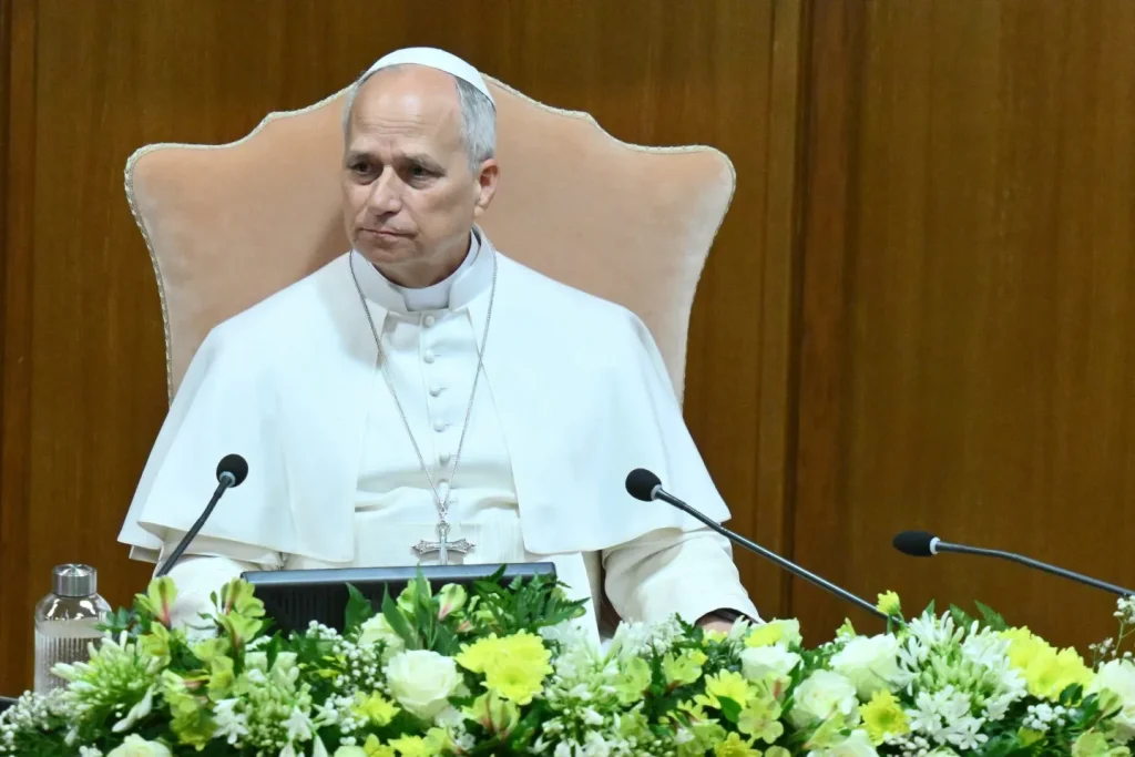 Religious leader in white robes sits at a desk with two microphones and a floral arrangement, wearing a cross necklace and a white skullcap.