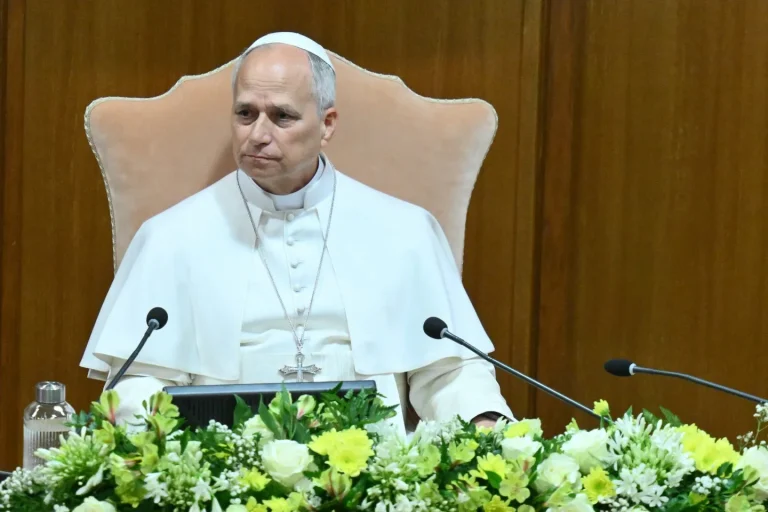 Religious leader in white robes sits at a desk with two microphones and a floral arrangement, wearing a cross necklace and a white skullcap.