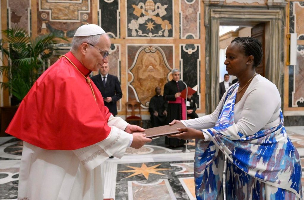Religious official in red robes hands a folder to a woman in a blue and white scarf during a formal ceremony in a marble hall.