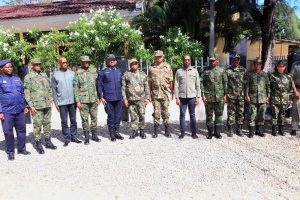 Group of soldiers in camouflage and blue uniforms standing in a line outdoors, with a gravel ground and greenery behind.