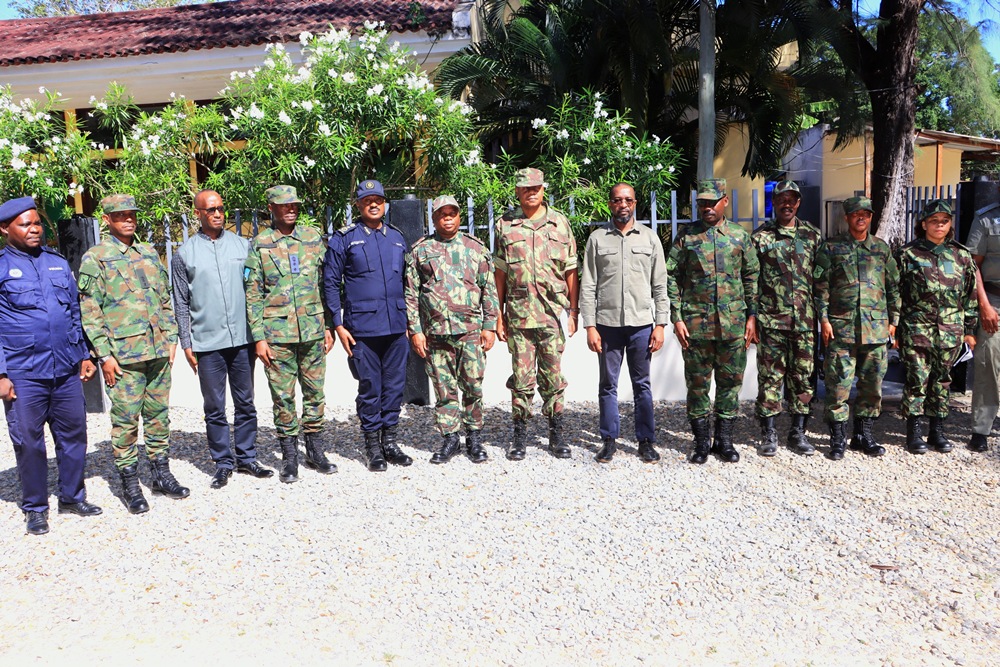 Group of soldiers in camouflage and blue uniforms standing in a line outdoors, with a gravel ground and greenery behind.