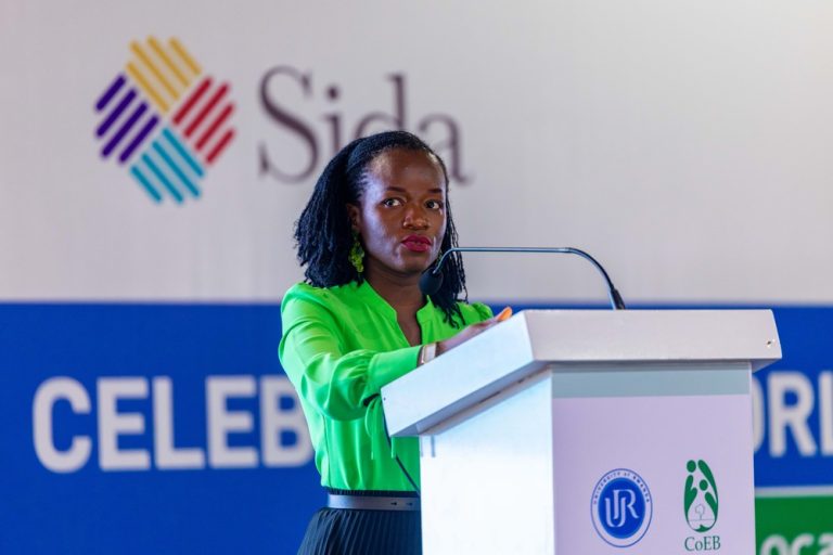 A woman in a neon green blouse stands at a white podium with a microphone, giving a speech against a backdrop with the Sida logo.