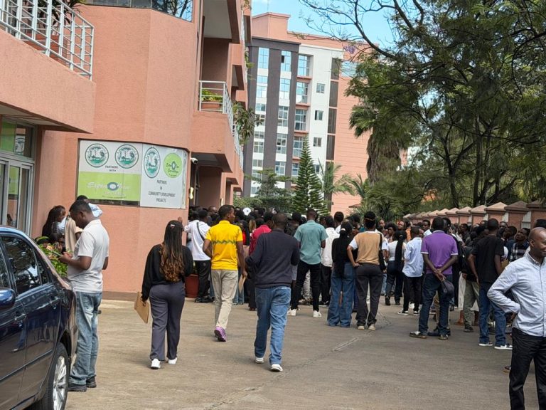 Crowd of people standing in line outside a peach-colored building with banners on the wall and trees nearby.