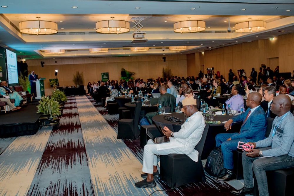 Audience seated at round tables in a conference hall, listening to a speaker at a podium on a stage.