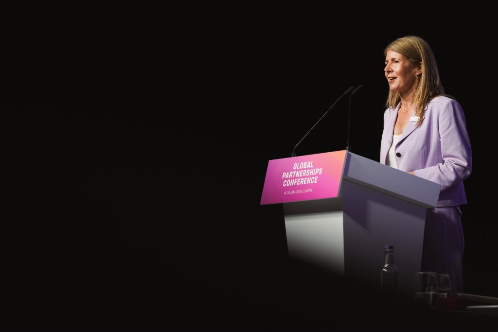 Woman in a lavender blazer speaks at a white podium with a pink conference sign reading 'Global Partnerships Conference' (London, May 19–20, 2026).