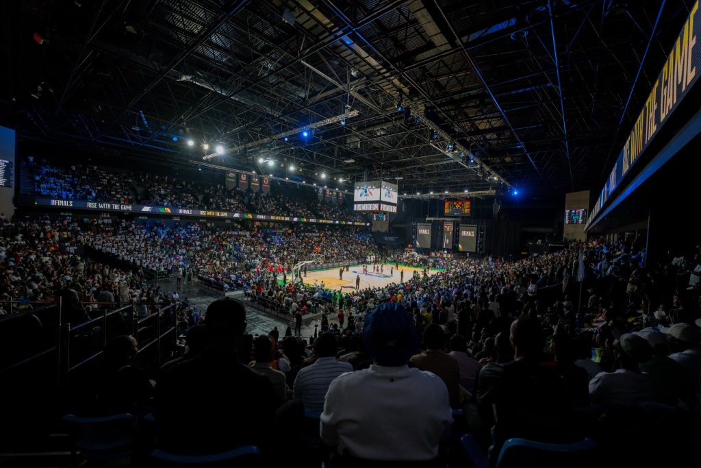 Indoor basketball arena filled with spectators watching a game, bright court under overhead rigging and screens above.