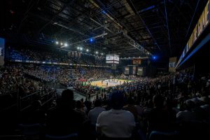 Indoor basketball arena filled with spectators watching a game, bright court under overhead rigging and screens above.
