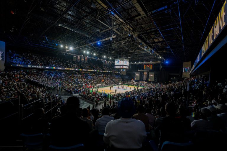 Indoor basketball arena filled with spectators watching a game, bright court under overhead rigging and screens above.