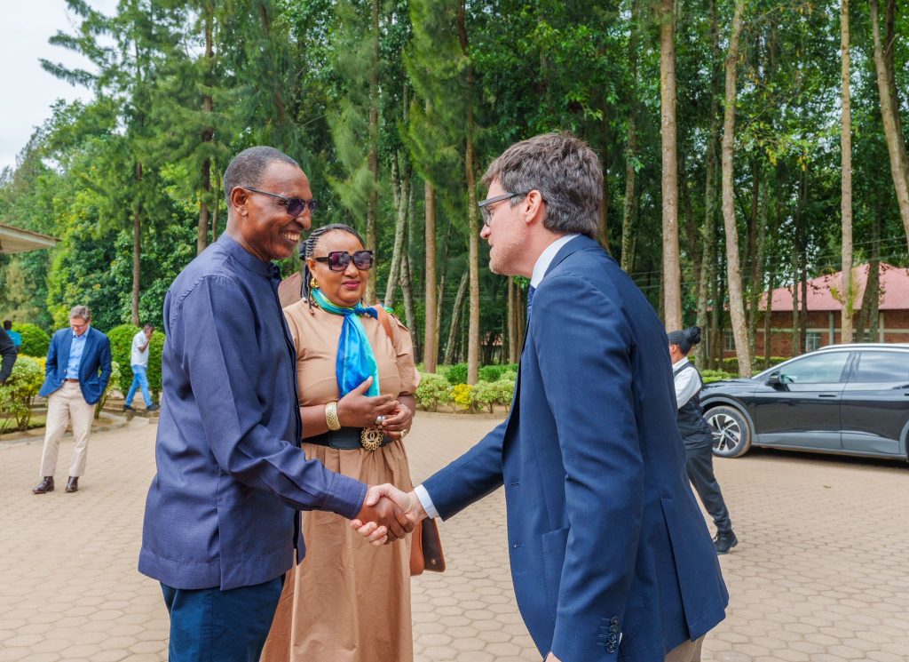 Three professionals shake hands outdoors; two men in suits and a woman in beige, with trees behind them in a courtyard setting.