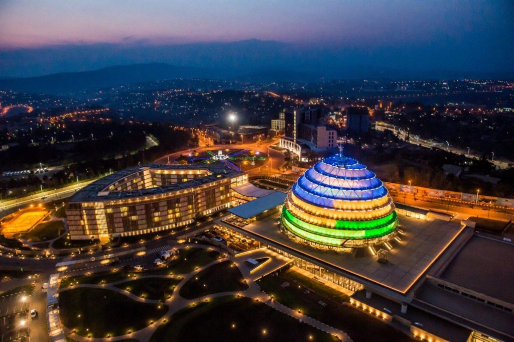 Aerial view of a circular, multi-colored illuminated dome building at dusk within a modern campus and surrounding city lights.