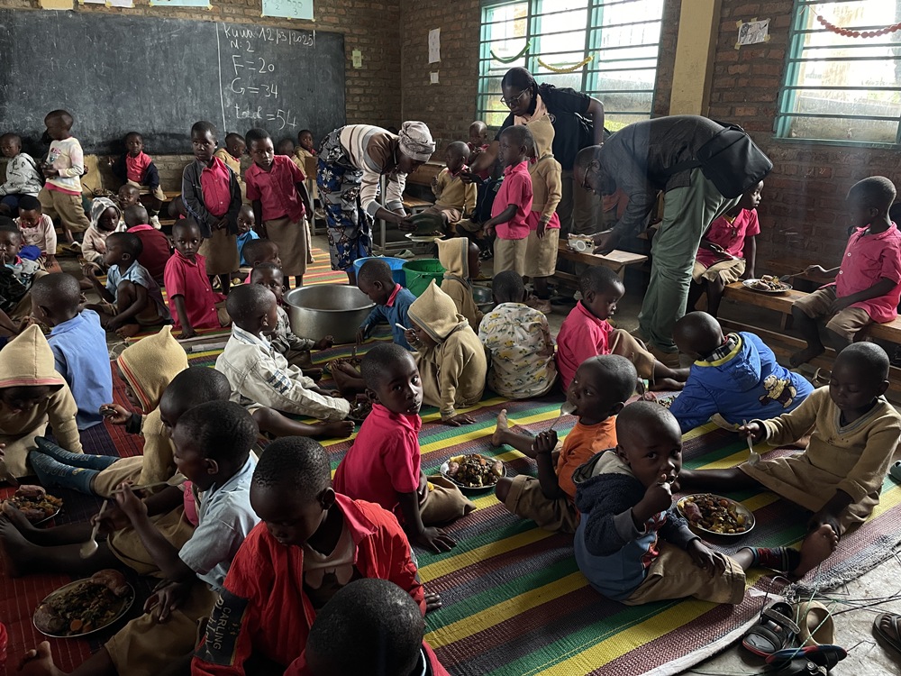 Group of children sitting and eating meals on a colorful rug in a classroom, as adults serve food nearby.