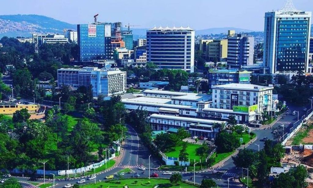 Aerial view of a modern city with tall glass buildings, green parks, and a curved multi-lane road intersection in the foreground.