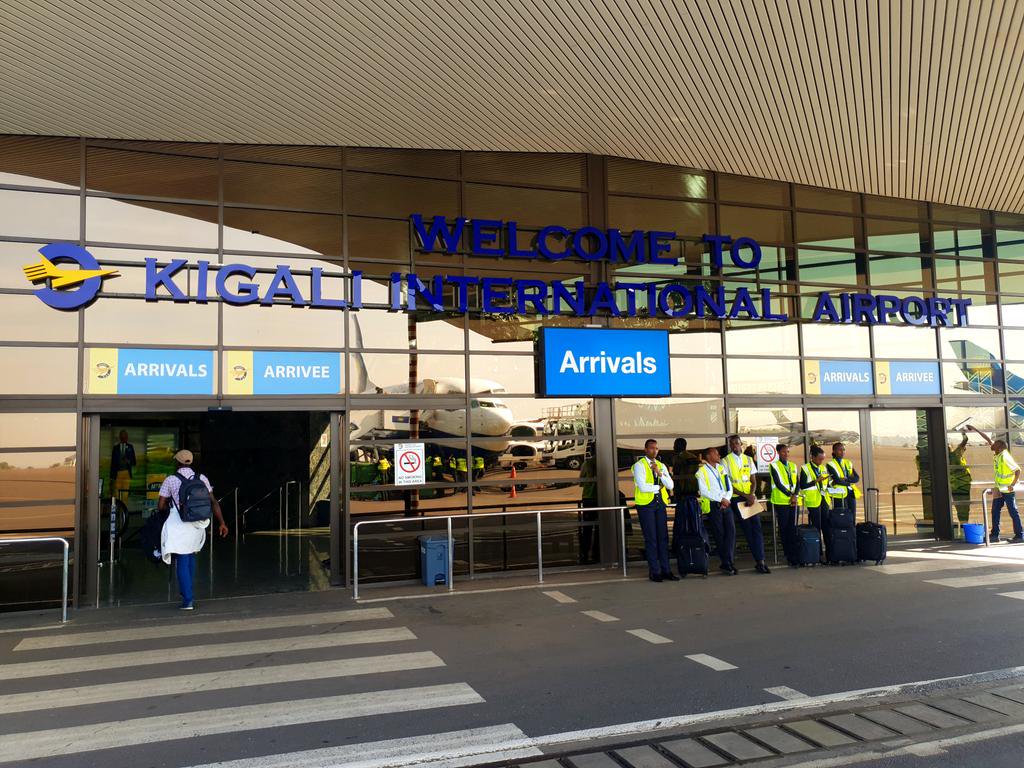 Front view of Kigali International Airport arrivals area with a blue 'Arrivals' sign and a line of uniformed staff beside a parked airplane.