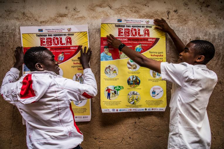 Two men put up bright yellow Ebola awareness posters on a rough wall.