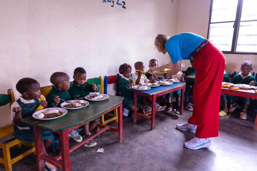 Adult in a blue shirt and red pants serves lunch to children at colorful tables in a classroom setting.