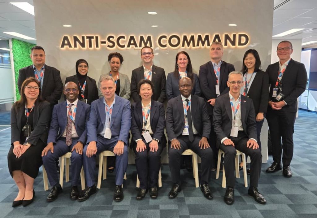 Group of diverse professionals in business attire posing for a photo in front of an 'Anti-Scam Command' sign in a modern office lobby.