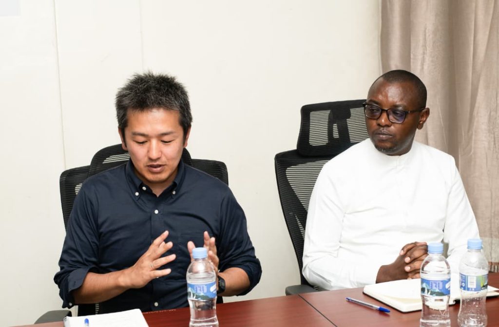Two men sit at a conference table; one speaks with expressive hand gestures while the other listens wearing white traditional attire and glasses, with water bottles on the table.