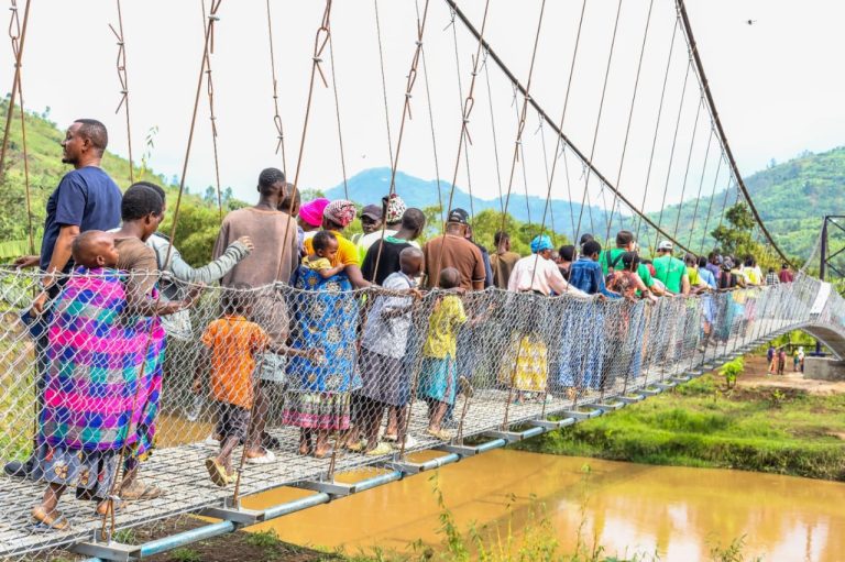 Crowd of people walking across a long suspension bridge over a muddy river in a rural valley with green hills in the background.