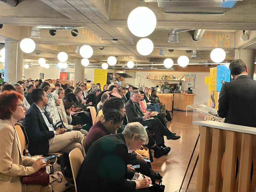 Audience seated in a modern venue listening to a speaker at a wooden podium, with round hanging lights overhead.