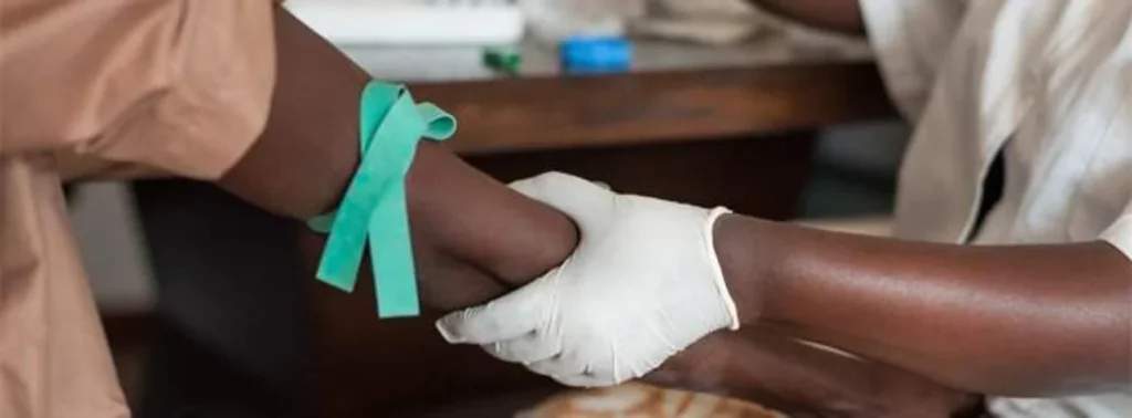 Caregiver wearing gloves supports a patient’s forearm as a teal band is tied around the wrist.