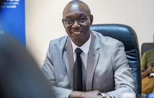 Smiling man in a light gray suit and dark tie sits at a desk in an office setting.