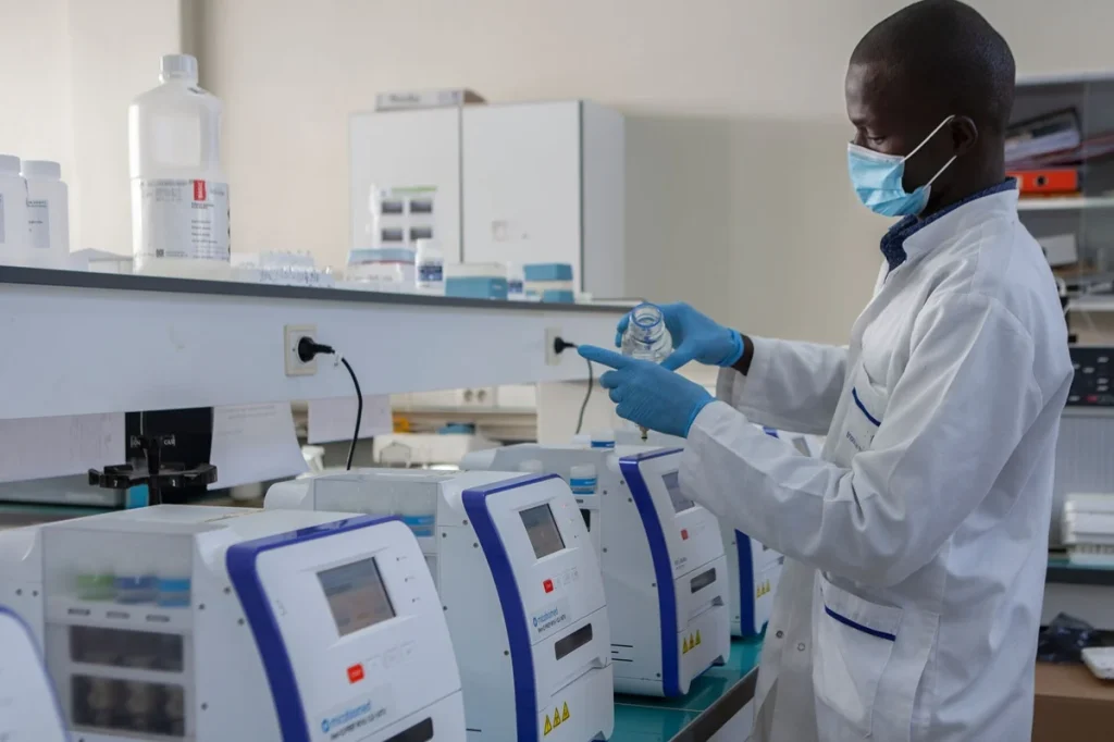 Lab technician in PPE (mask and gloves) handling a bottle at a clinical lab bench with multiple automated devices nearby.
