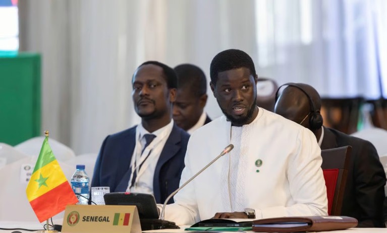 Senegalese delegate in white traditional attire speaks into a microphone at a formal conference, with a Senegal flag and SENEGAL nameplate on the table.