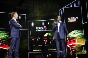 Two men in dark suits speak into microphones on a stage with 'Africa Forward' visuals behind them.