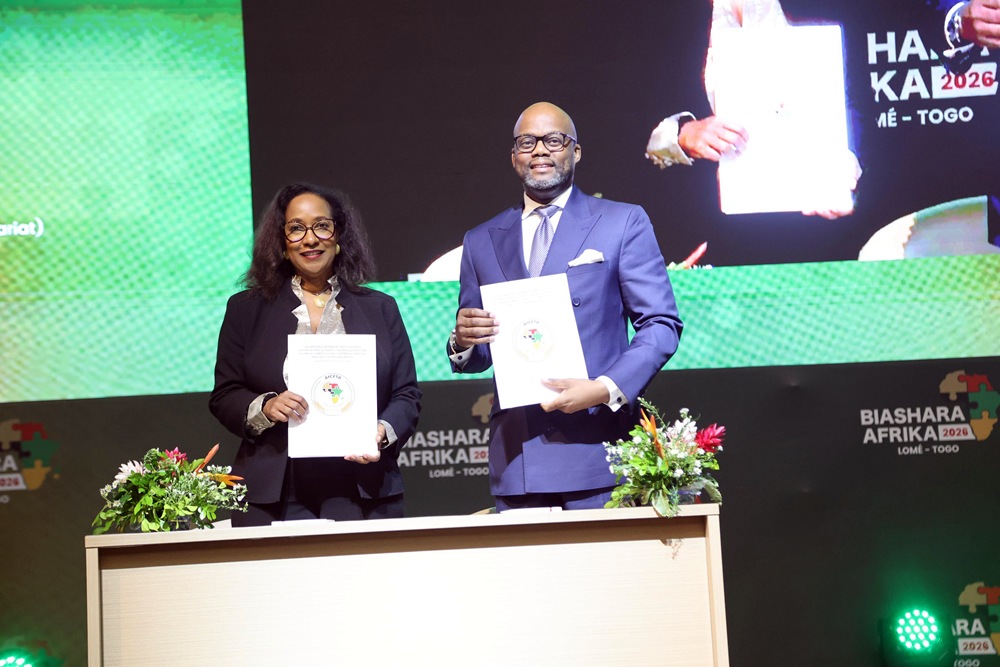 Two professionally dressed individuals stand behind a desk on a stage, each holding a document and smiling during a formal signing ceremony.