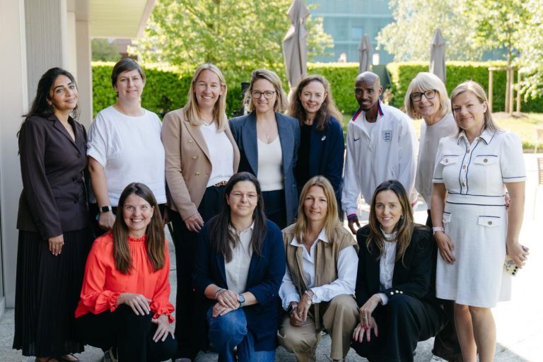 A diverse group of professionals, mostly women, posing outdoors in smart casual attire with smiles on a sunny day.