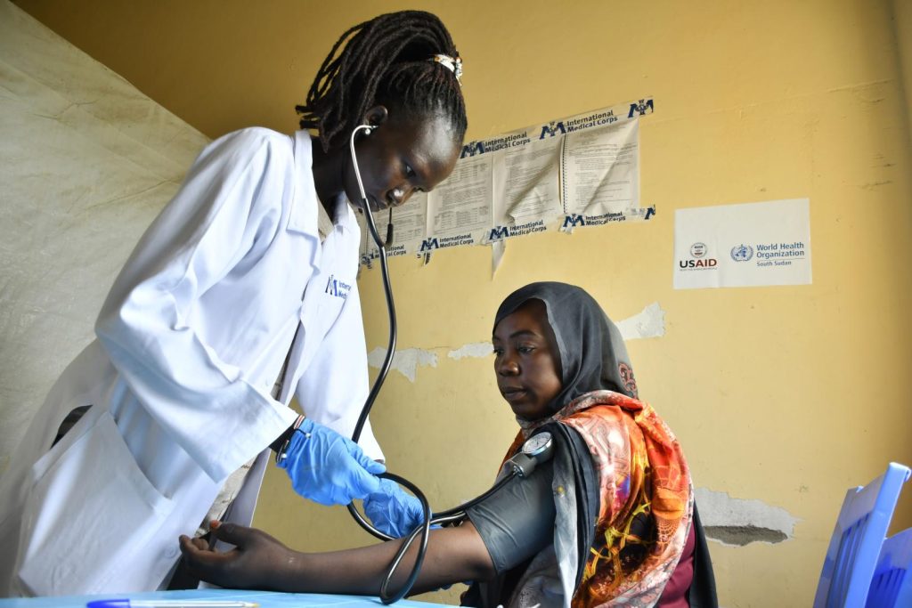 Healthcare worker in a white coat using a stethoscope to measure a woman's blood pressure in a clinic.