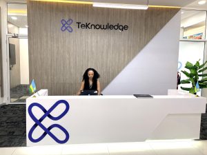 Reception desk with a woman at a laptop, TekKnowledge logo on a wooden wall behind, and small flags on the counter.
